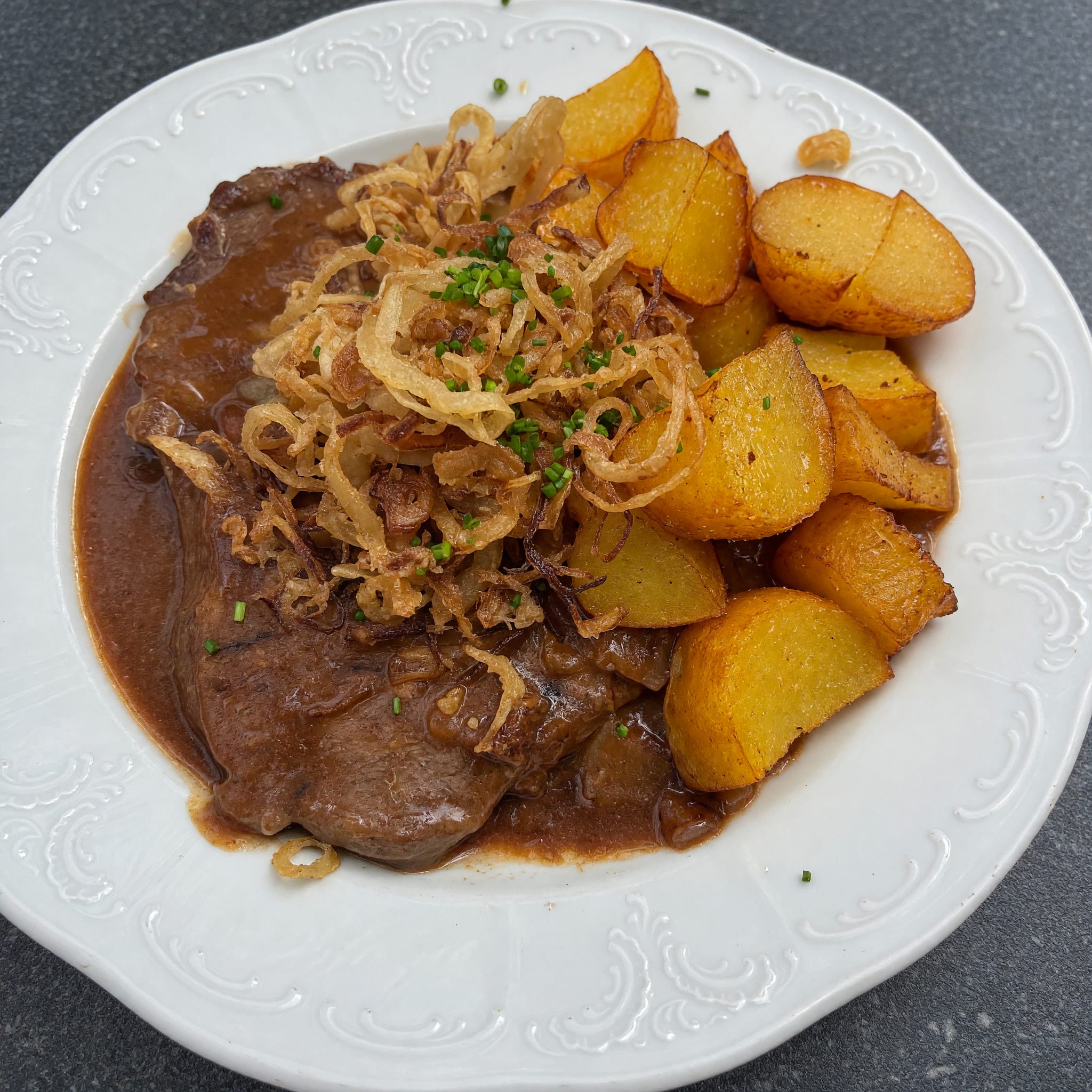 Rostbraten mit Röstzwiebeln und Braterdäpfeln auf einem weißen Teller mit Reliefrand, fotografiert von einem Gast