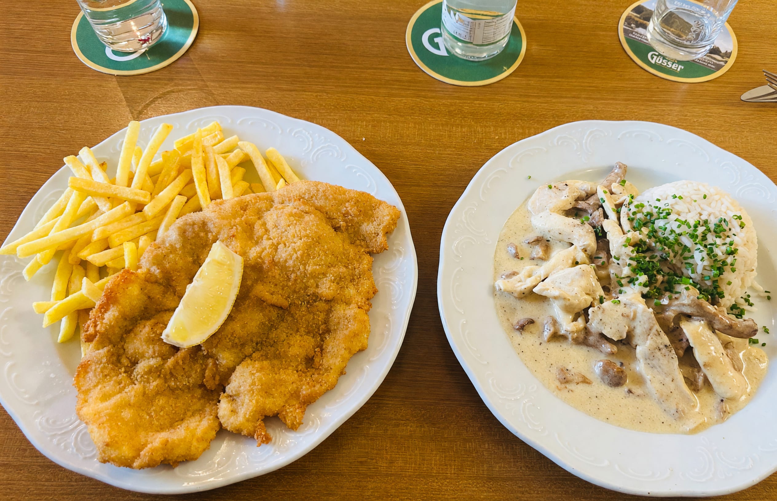 Wirtshaustisch von oben: ein Schnitzel mit Pommes frites und Zitrone, daneben ein Rahmgericht mit Reis, Gläser auf Bierdeckeln
